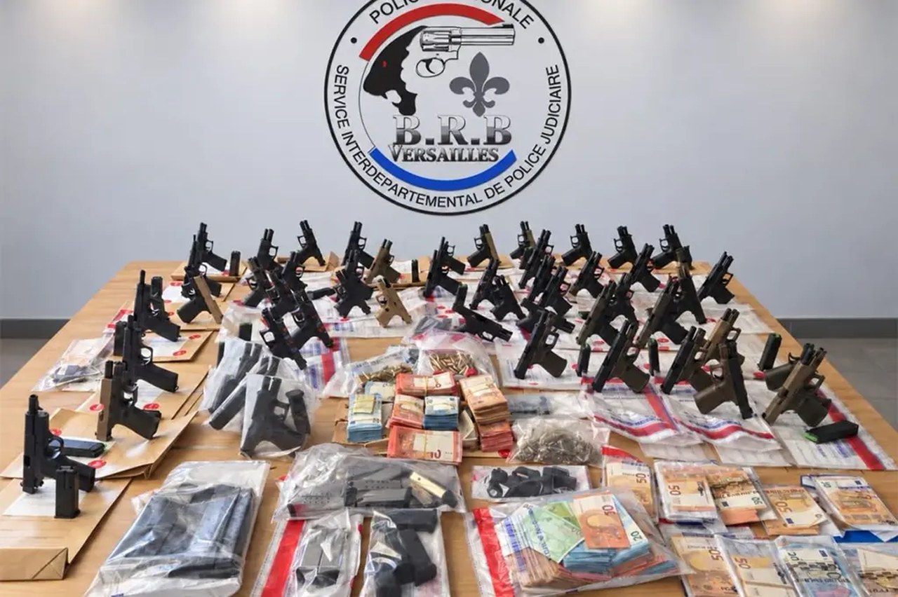 Dizaines de pistolets neufs, chargeurs et liasses de billets exposés sur une table devant l'écusson de la BRB de Versailles